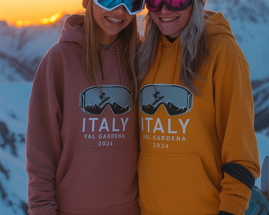 Two smiling women wearing ski goggles and hoodies with a snowboarder logo and the text ‘Italy Val Gardena 2024,’ standing together in snowy mountains during a vibrant sunset.